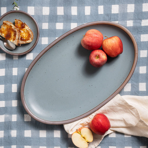 Three red apples on a large ceramic oval platter in a blue-grey color with unglazed rim, sitting on a crosshatch blue-grey tablecloth.