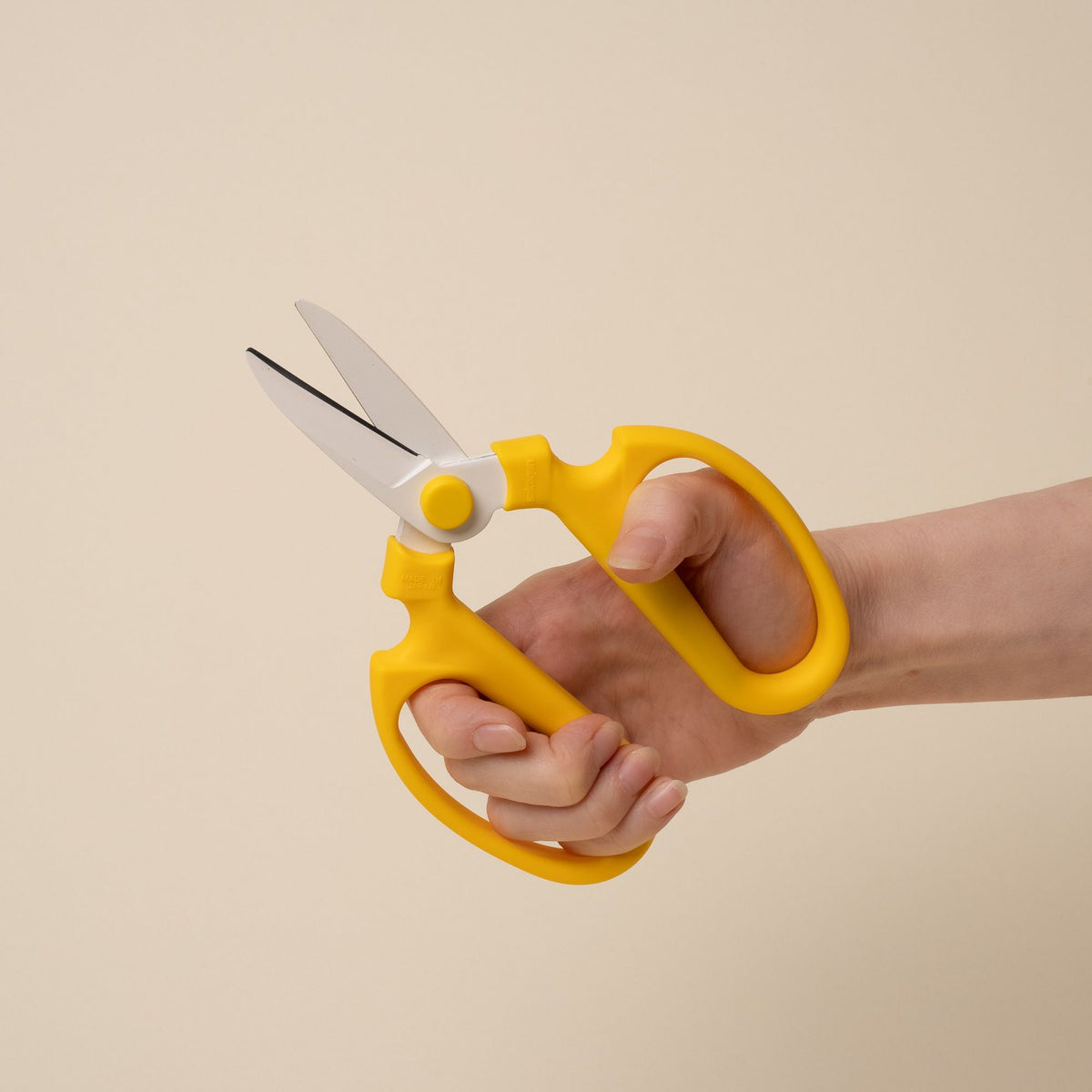 A hand holds an open pair of flower scissors with large yellow handles and short blades.