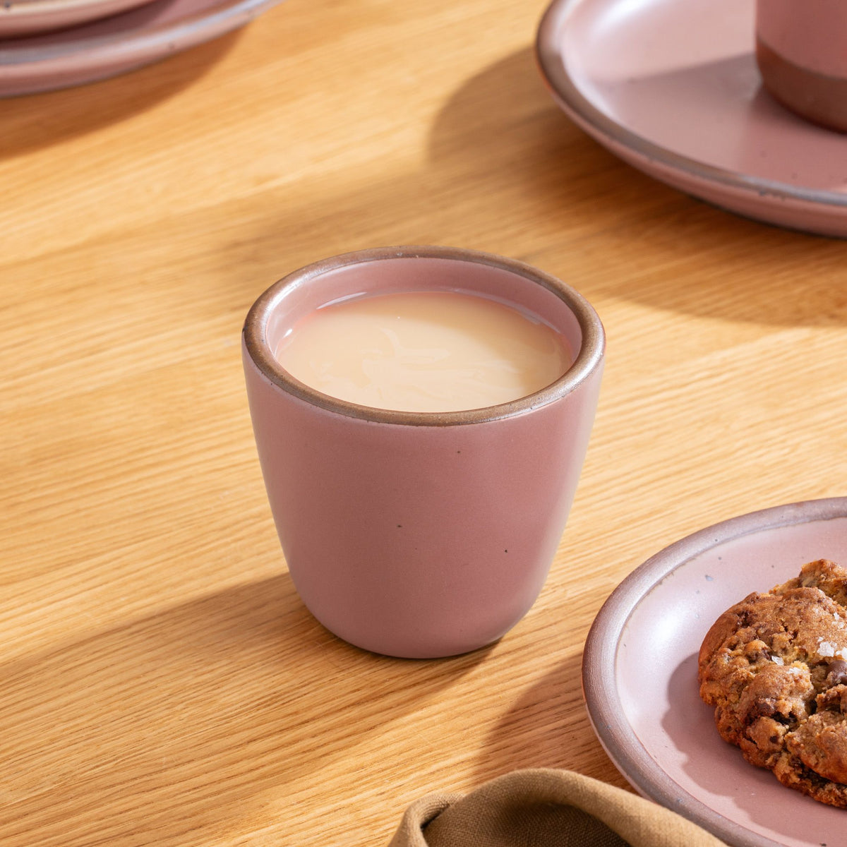 Dusty mauve pink ceramic cup filled with milk tea or coffee, next to a cookie on a matching plate atop a wooden table.