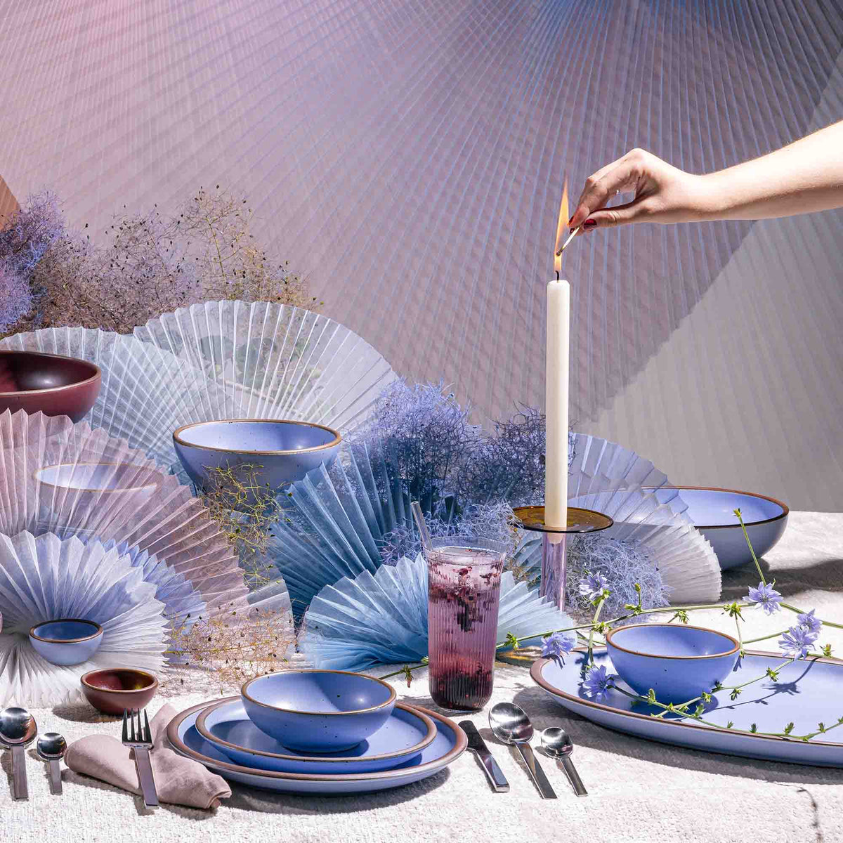 A hand lights a white taper candle in a beautiful tablescape featuring artful fabrics, periwinkle dishes, and silverware.