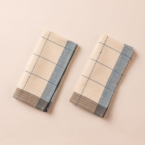 Two folded natural colored napkins side by side that are designed with turquoise gridlines with light blue and tan striped edging.
