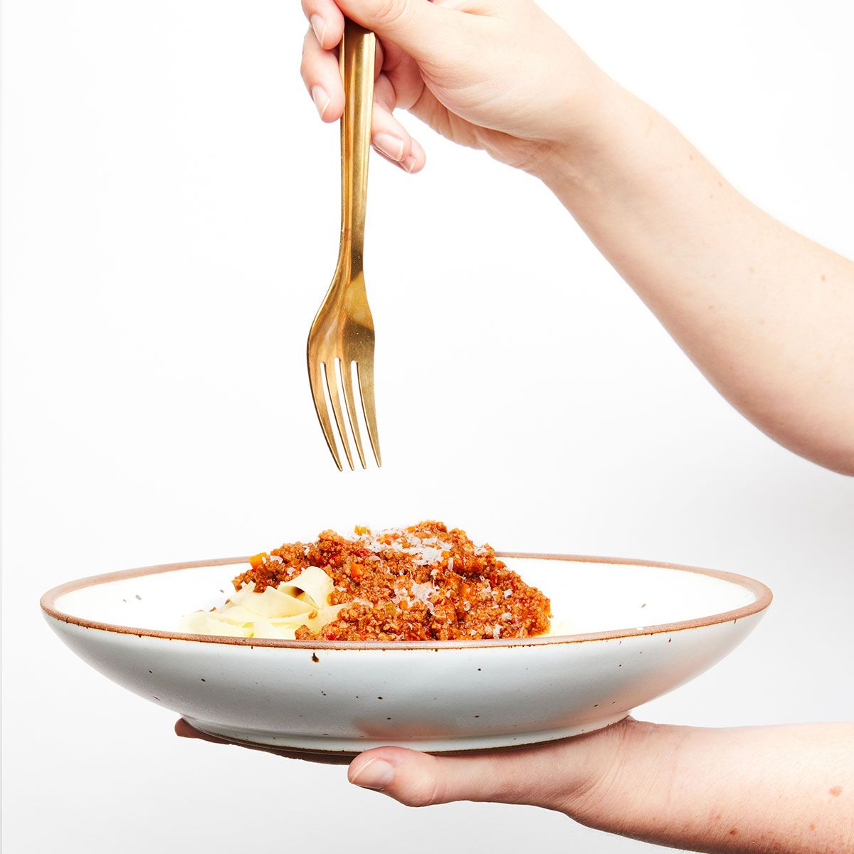 A hand holds a brass fork over a white ceramic coupe plate filled with pasta.