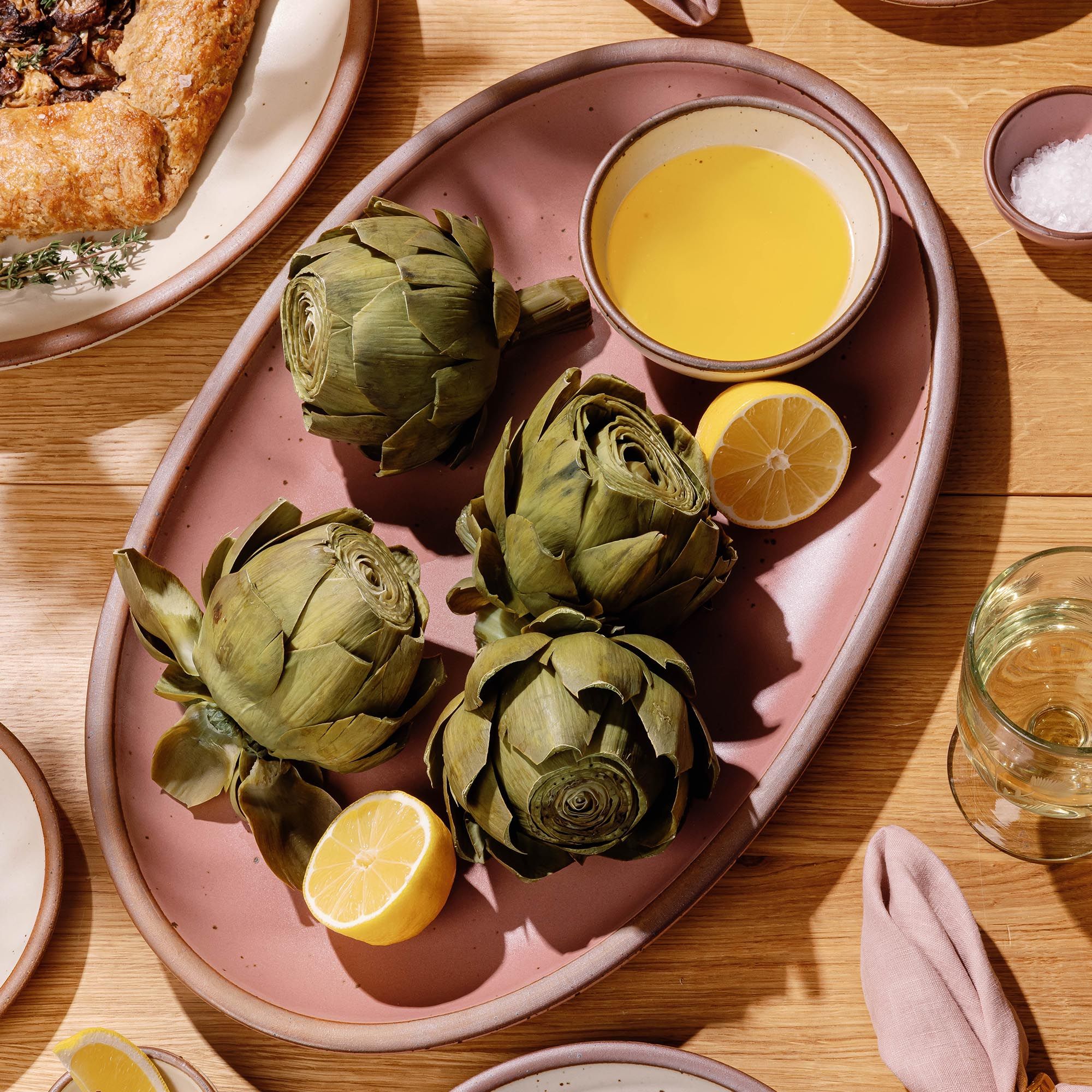 Artichokes on a dusty mauve pink oval platter with lemon halves and a bowl of melted butter on a wooden table.