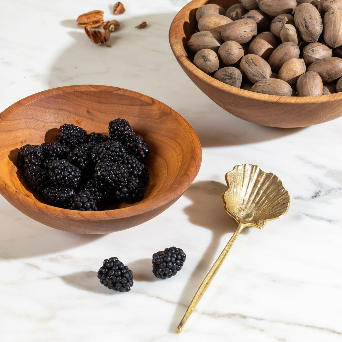 Wooden bowls filled with blackberries and pecans on marble, with a gold shell-shaped spoon.