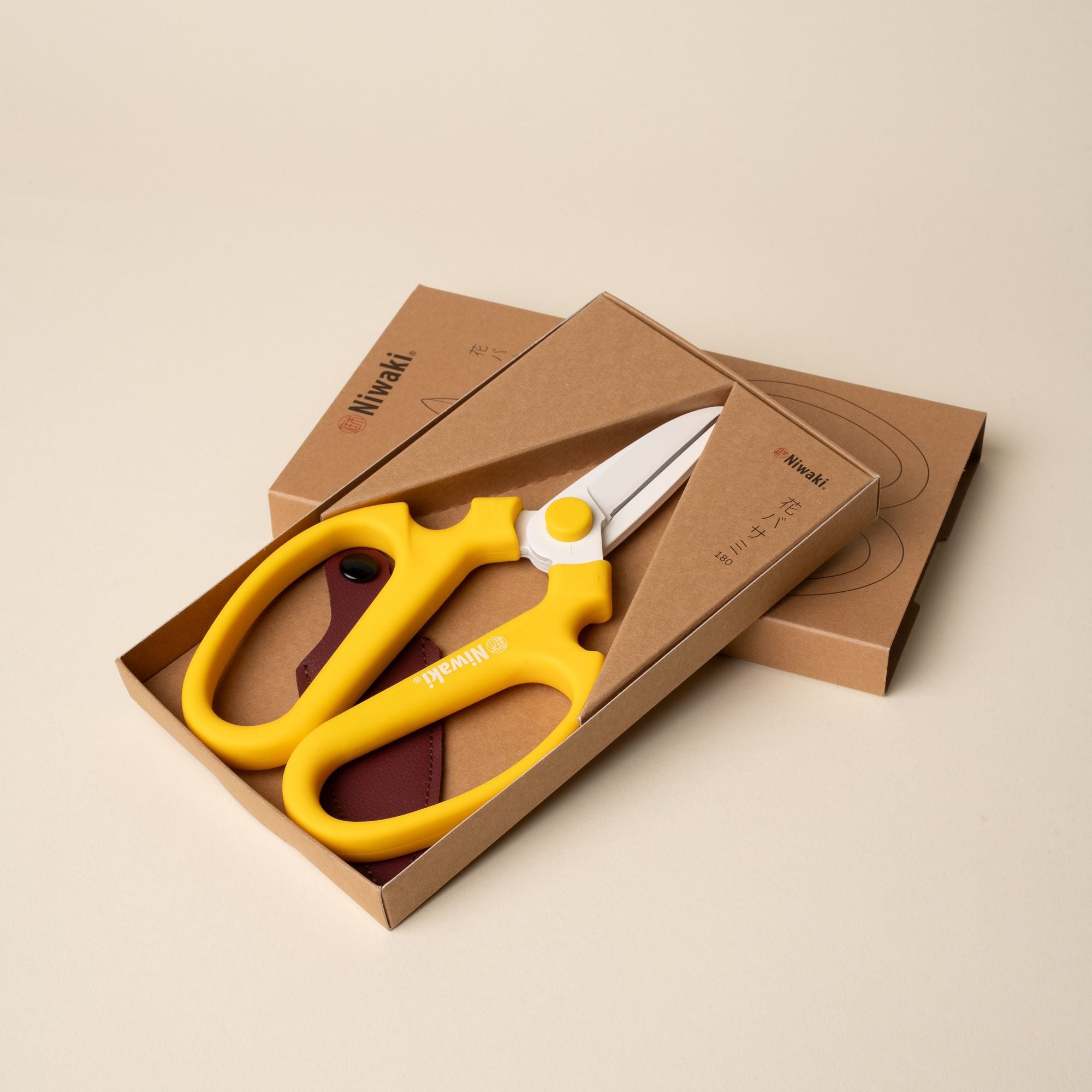 A pair of flower scissors with large yellow handles and short blades sitting in a kraft packaging box.