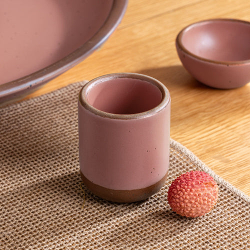 Small dusty pink ceramic cup on a woven placemat with a lychee fruit beside it on a wooden table.