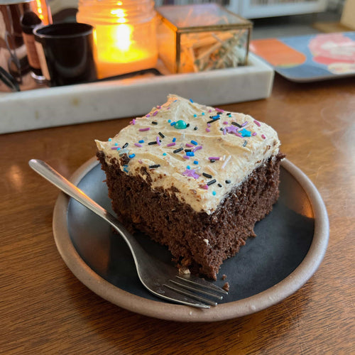 A slice of chocolate cake with cream frosting and sprinkles sits on a black ceramic cake plate with a fork.