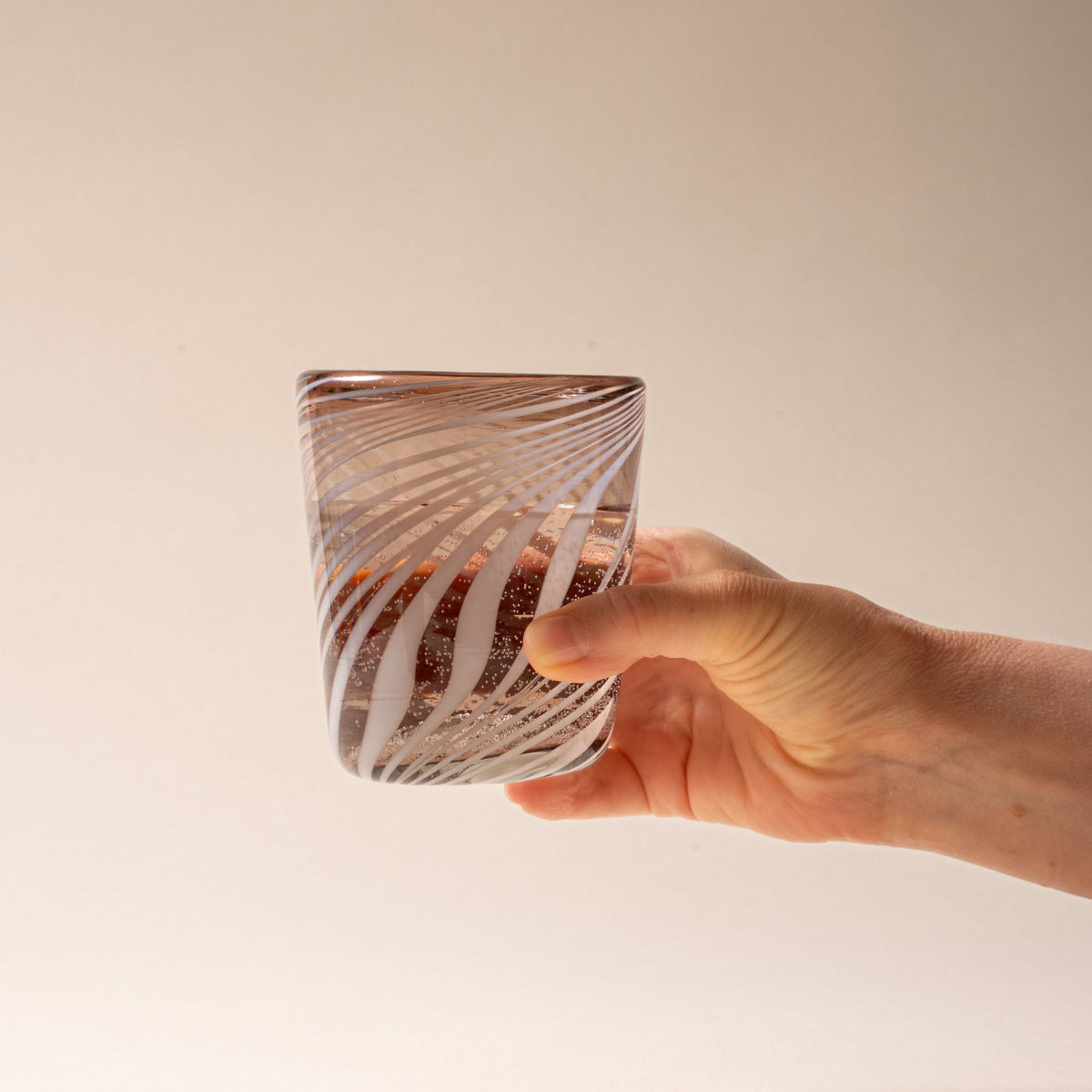 Hand holds out a smoky brown glass tumbler with white diagonal swirl pattern, filled with water, on a neutral background.