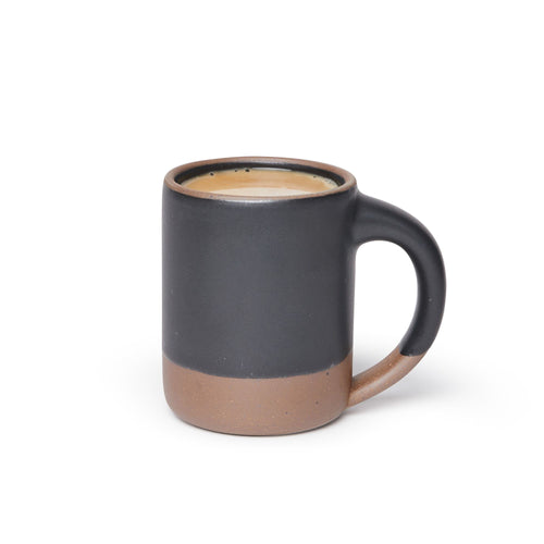 A big sized ceramic mug filled with coffee with handle in a graphite black glaze featuring iron speckles and unglazed rim and bottom base.