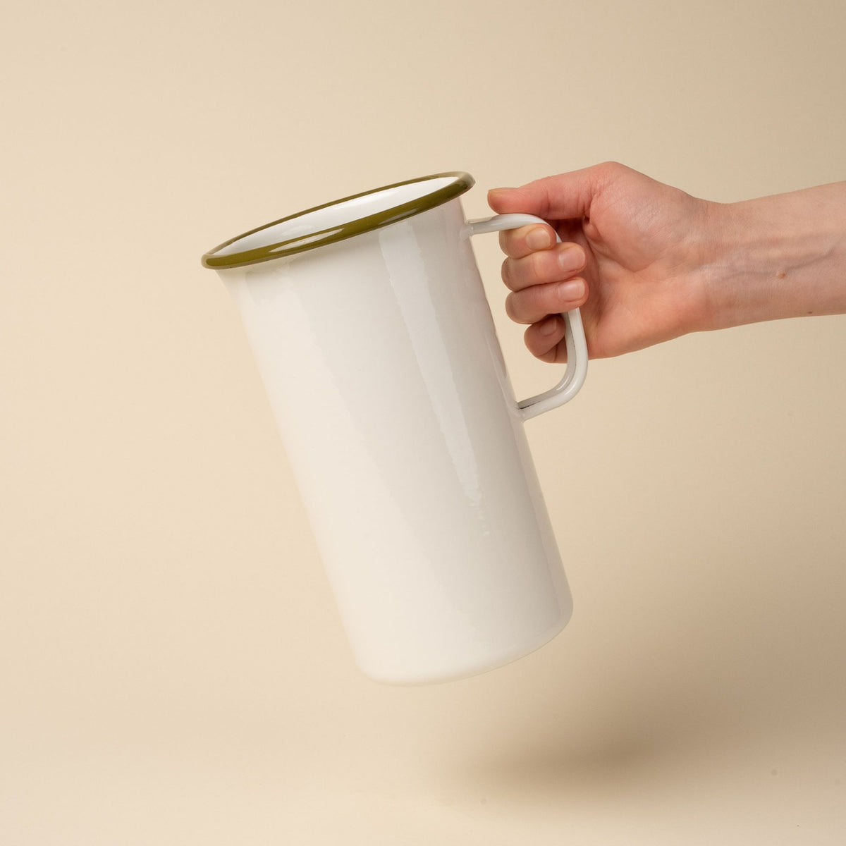 A hand holds out a large enamel pitcher with handle in a white color with an olive green rim.