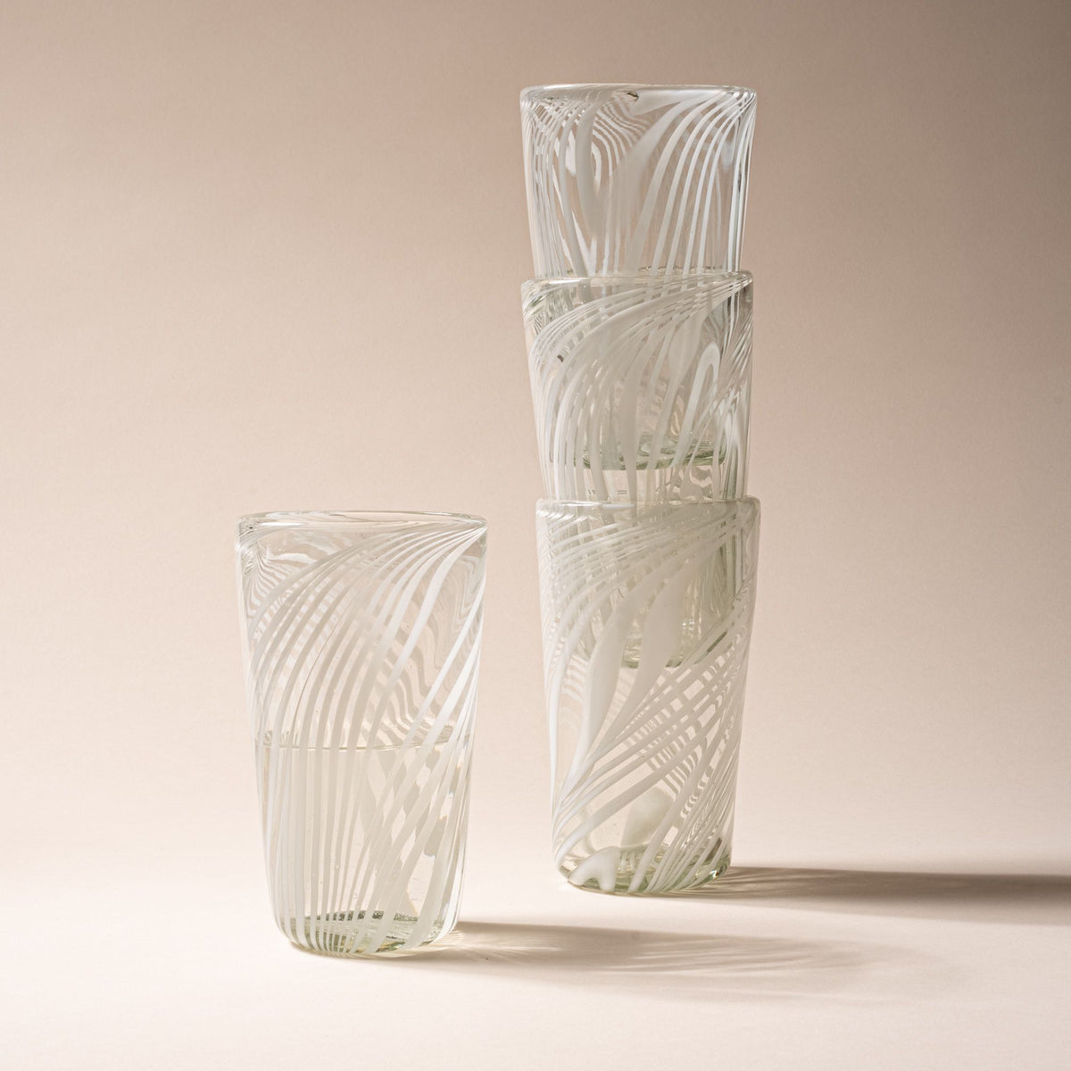 Set of four clear glass tumblers with white swirling stripe design, three stacked and one standing beside on neutral backdrop.