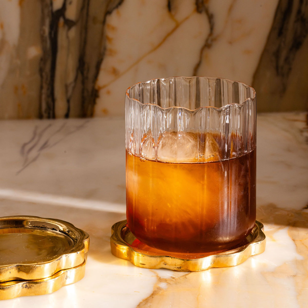 Amber cocktail with ice in a ribbed glass, set on a gold coaster against a marble background.