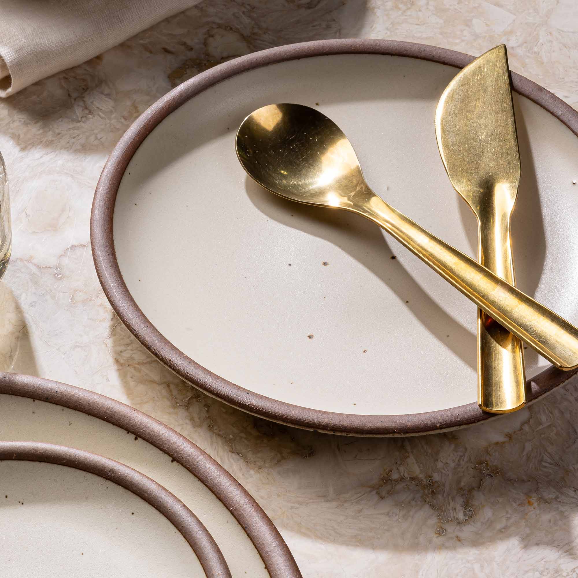 Ceramic cream plate with unglazed rim with brass flatware sitting on top.