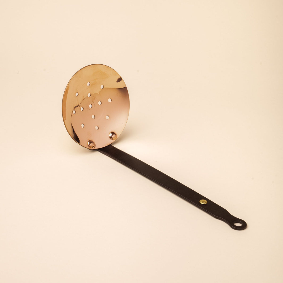 Round copper skimmer with perforated bowl and long black handle featuring a brass rivet and hanging loop, set on beige background.