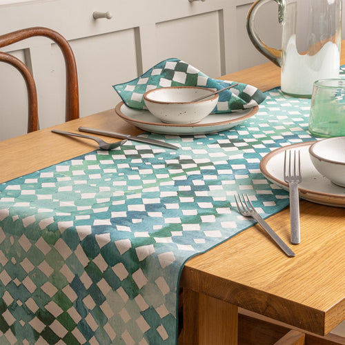 A wooden dining table styled with a teal patterned table runner, ceramic plates and bowls, silver utensils, and green glassware.