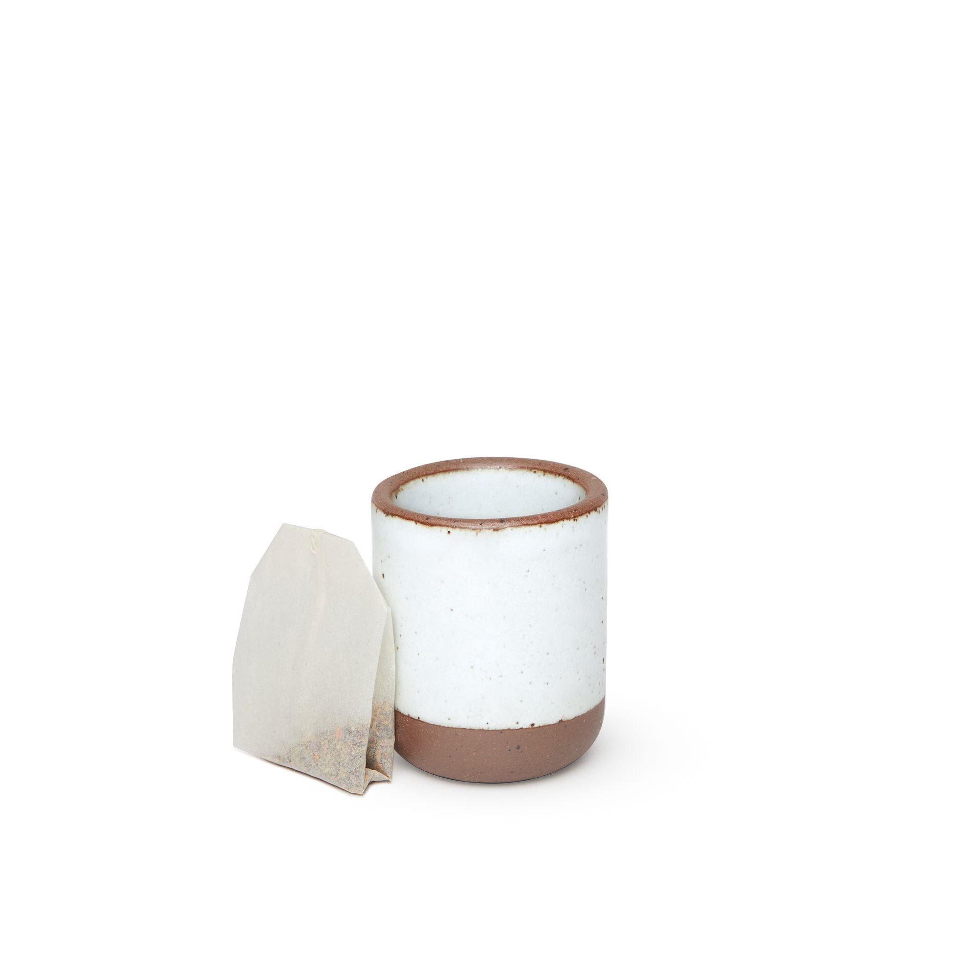 A teabag sitting next to a small, short ceramic mug cup in a cool white color featuring iron speckles and unglazed rim and bottom base.