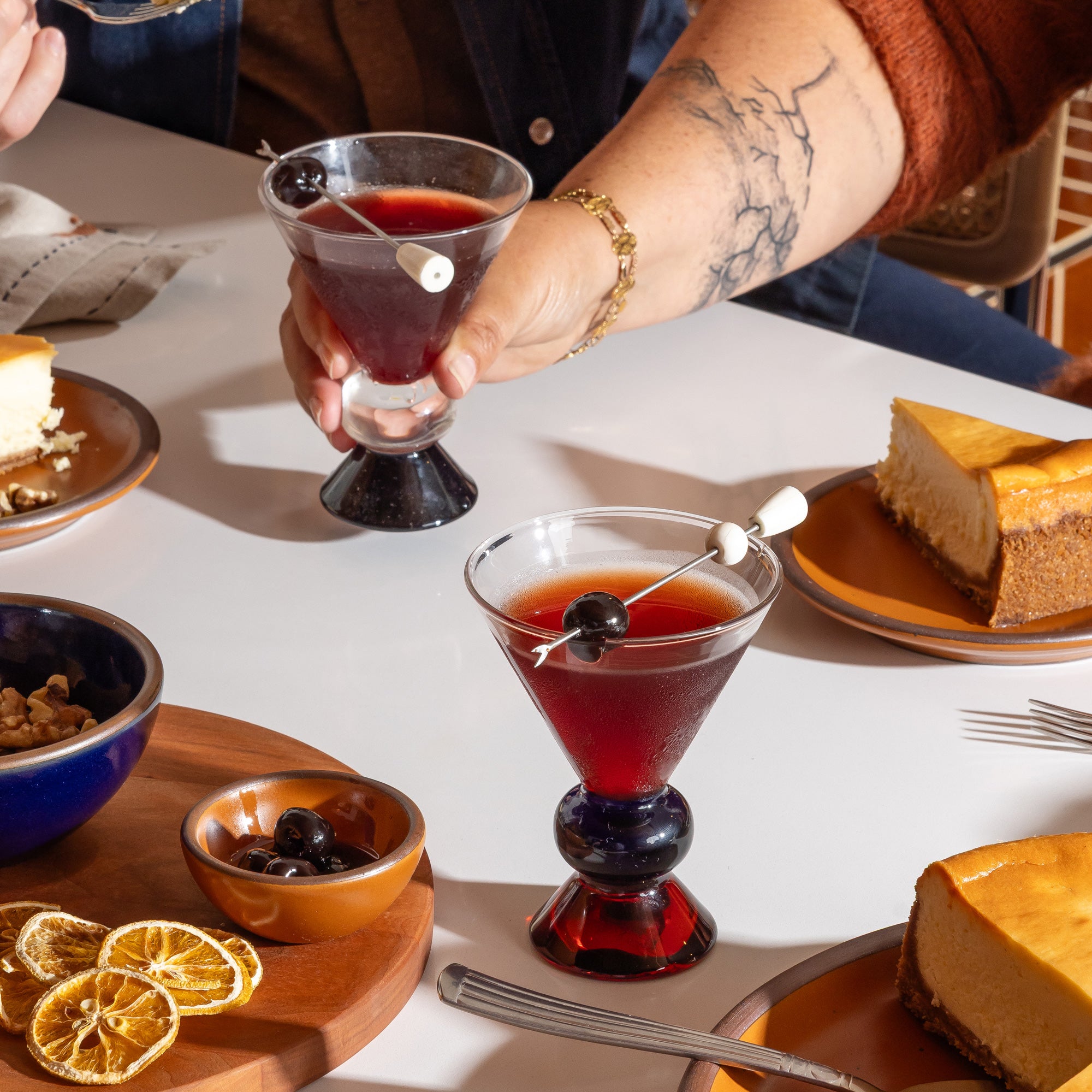 Two clear cocktail-style glasses with sculptural clear, blue and amber base, on a table with dessert and a hand holding one of the glasses.
