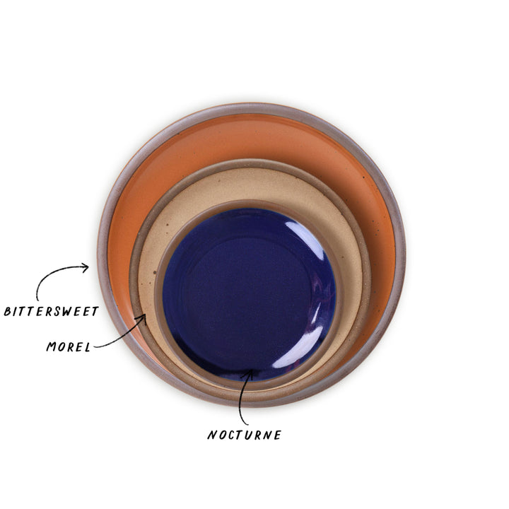 Three nested ceramic plates shown from a top-down view on a white background, with handwritten labels pointing to each: BITTERSWEET in the outer EARTHY ORANGE-toned plate, MOREL in the ligh brown plate, and NOCTURNE in the glossy dark blue plate.