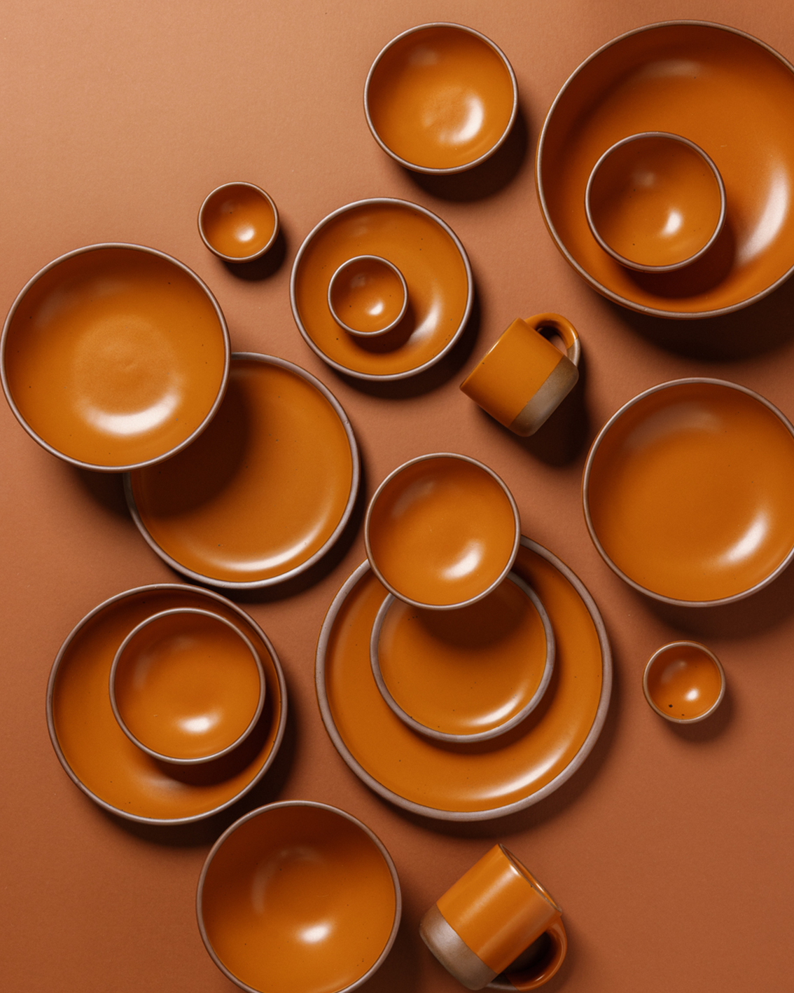 A coordinated set of matte earthy orange ceramic tableware arranged in a flat lay on a matching terracotta background, including plates, bowls, cups, and small dishes.