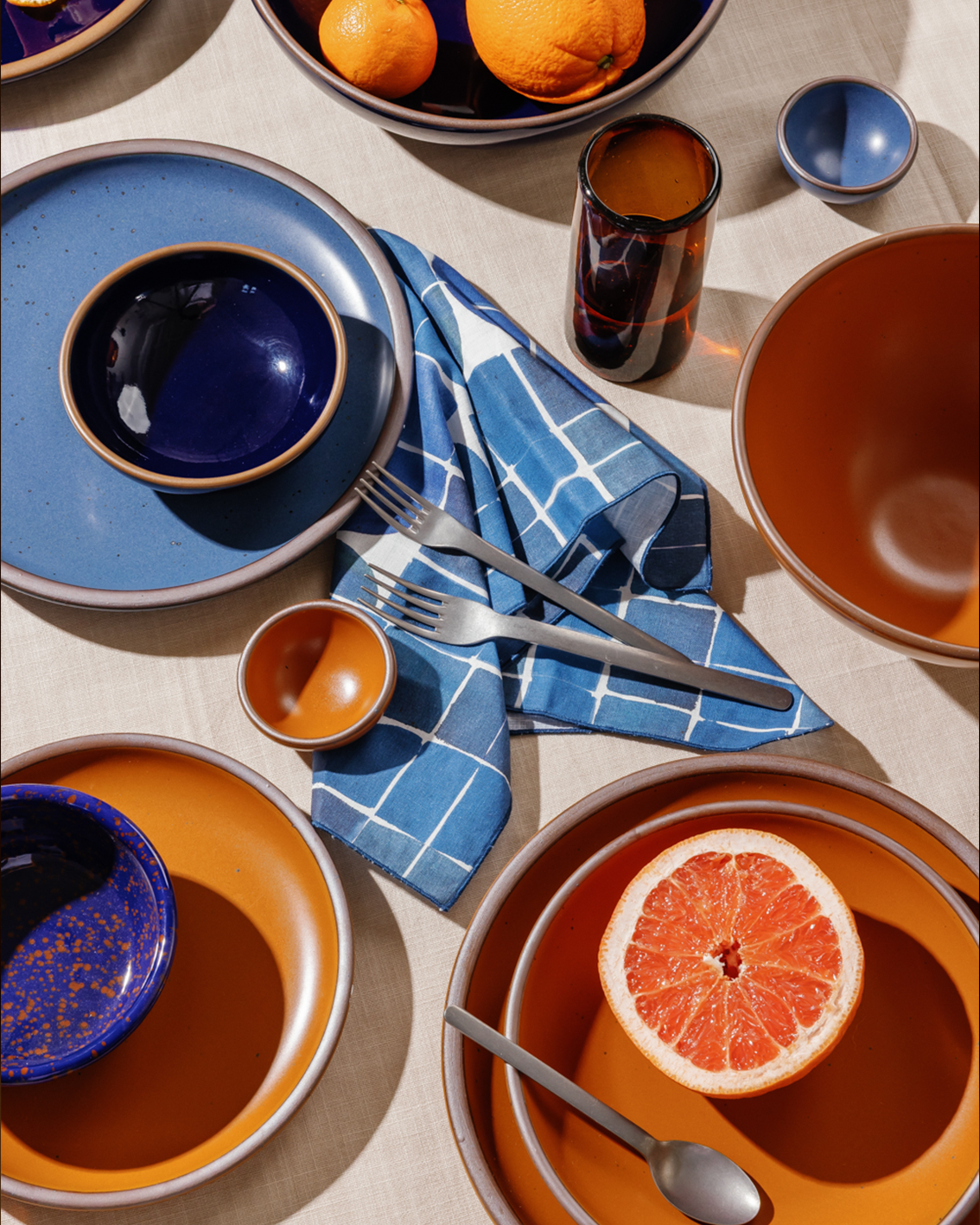 A styled tabletop flat lay of ceramic tableware in earthy orange and deep glossy blue, arranged with steel cutlery, a dark amber glass, a blue checkered napkin, and citrus fruit on a beige fabric surface.