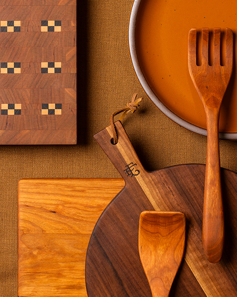 A styled flat-lay of wooden kitchen and serving tools on a warm brown textured surface, including a round wooden cutting board with a spoon and spatula, a rectangular cutting board with a checkerboard pattern, and a earthy orange plate with a wooden fork.