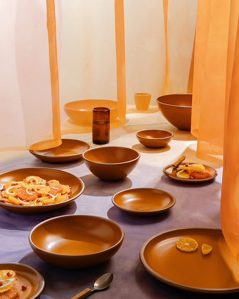 A set of matte earthy orange colored ceramic tableware, including plates, bowls, and a smoky amber tumbler glass, arranged on a table draped with a lavender cloth and sheer orange curtains. Citrus is styled on some plates as props.