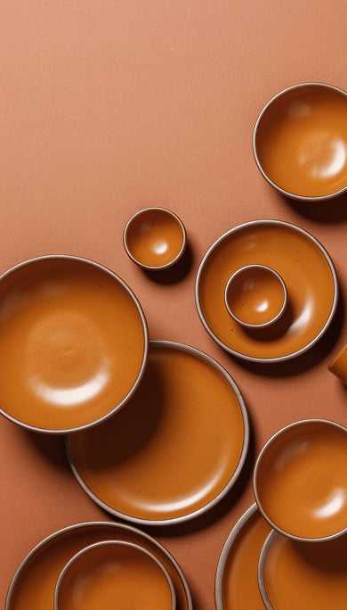 A set of matte earthy orange-colored ceramic tableware, including plates, bowls, and small cups, arranged in a flat-lay composition on a terracotta background.