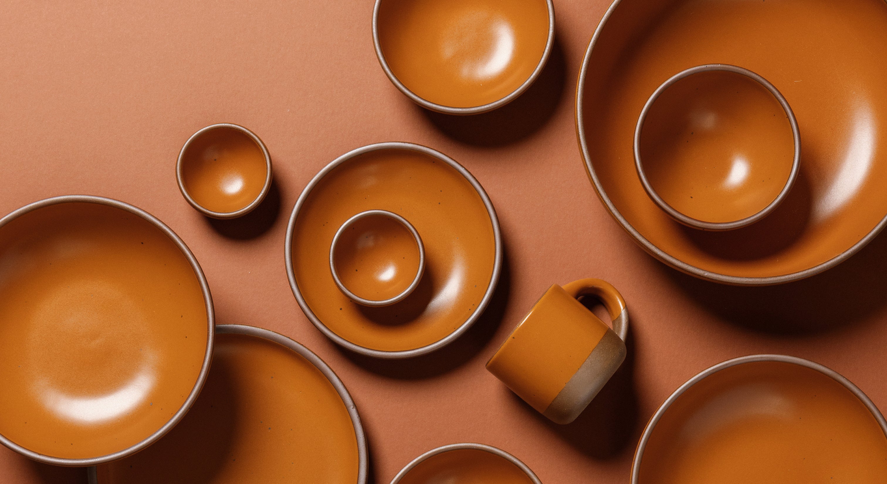A set of earthy orange-colored ceramic tableware, including bowls, plates, a mug, and small cups, arranged in a flat-lay composition on a terracotta background.
