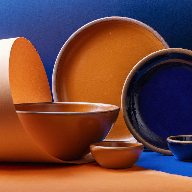 A styled studio arrangement of ceramic tableware in two contrasting glazes: a dark glossy blue and a warm earthy orange. The set includes plates, bowls, and small cups, some nested together and others displayed upright, set against an blue paper background and an orange paper loop. 