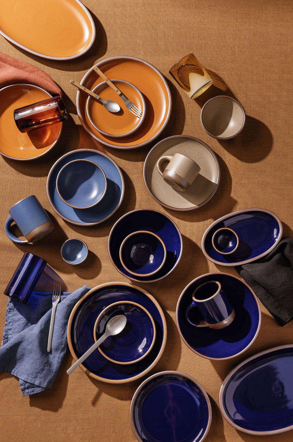 A styled flat-lay of ceramic tableware in glossy dark blue, earthy orange, and warm beige tones, arranged on a textured tan surface with matching napkins and cutlery.