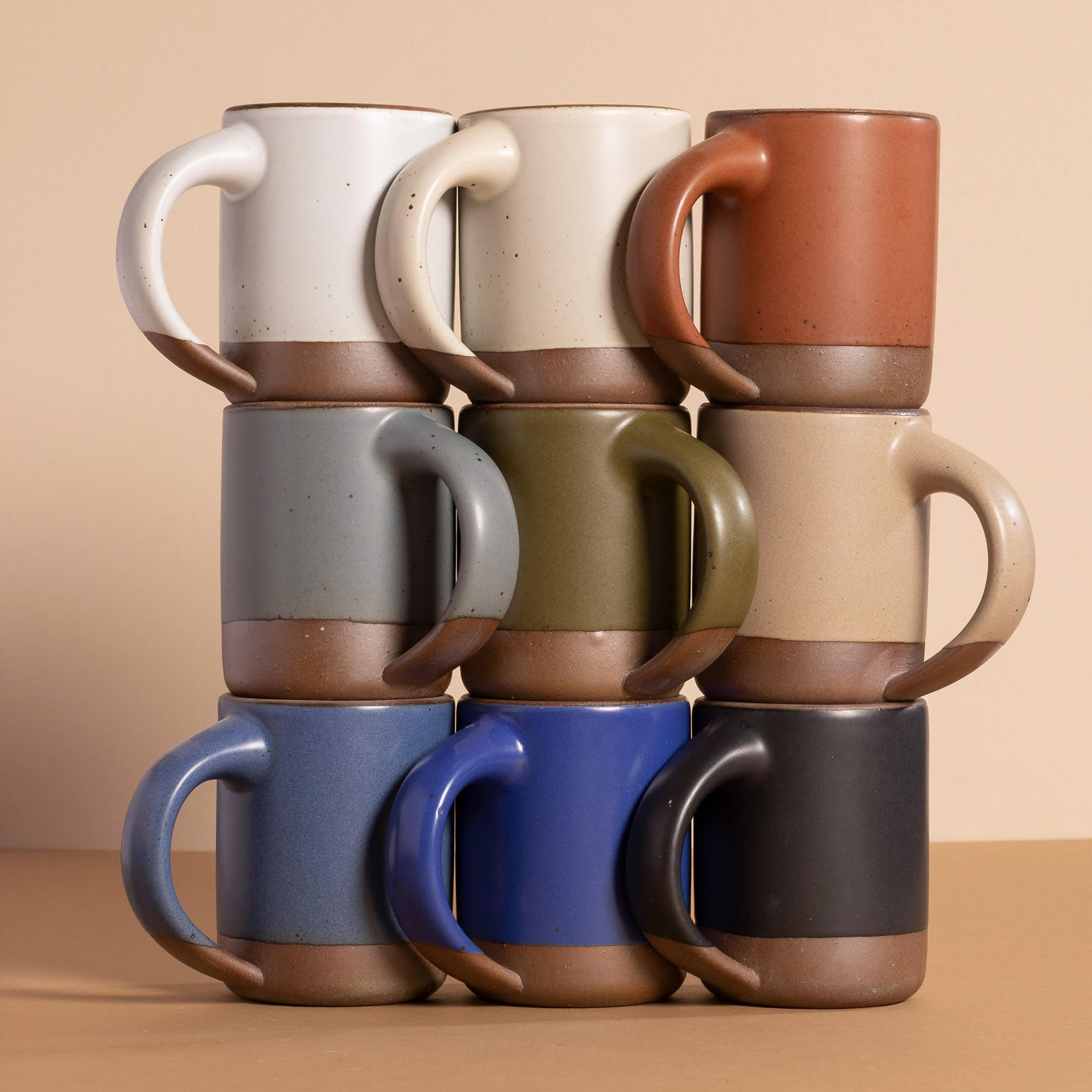 A stack of nine ceramic mugs with wide handles, arranged in a 3x3 grid. Each mug is glazed in a different color with and an unglazed base.
