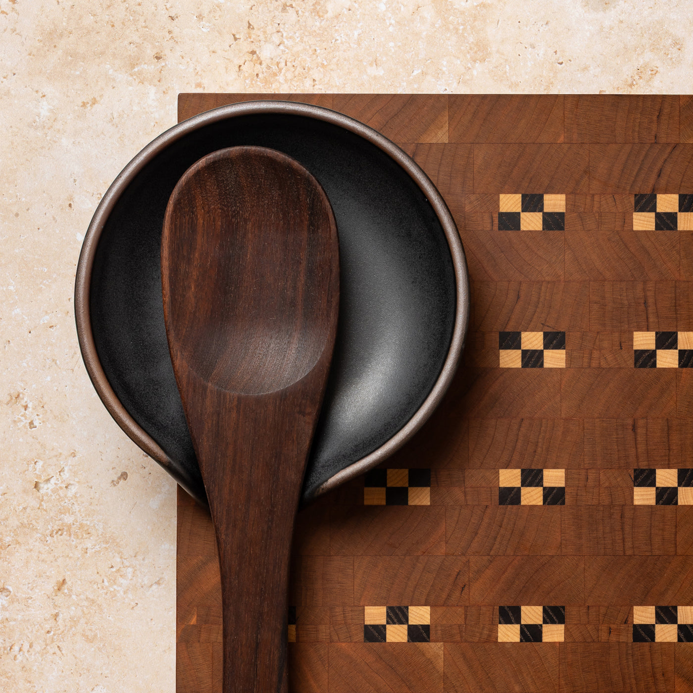 A wooden spoon sitting on a ceramic black graphite spoon rest on a quilted patterned cutting board.