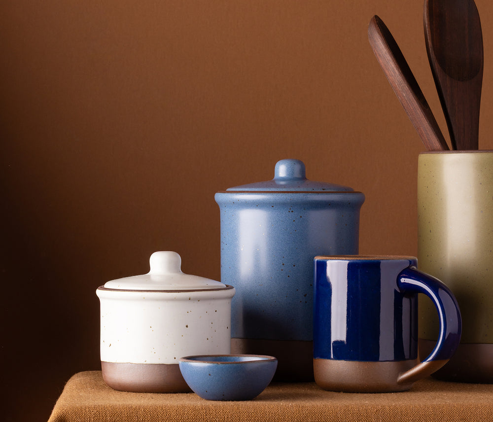 A collection of ceramic kitchen tabletop items such as a small jar, tall jar, utensil crock, along with a mug and bitty bowl, in neutral, blue, and olive green colors.