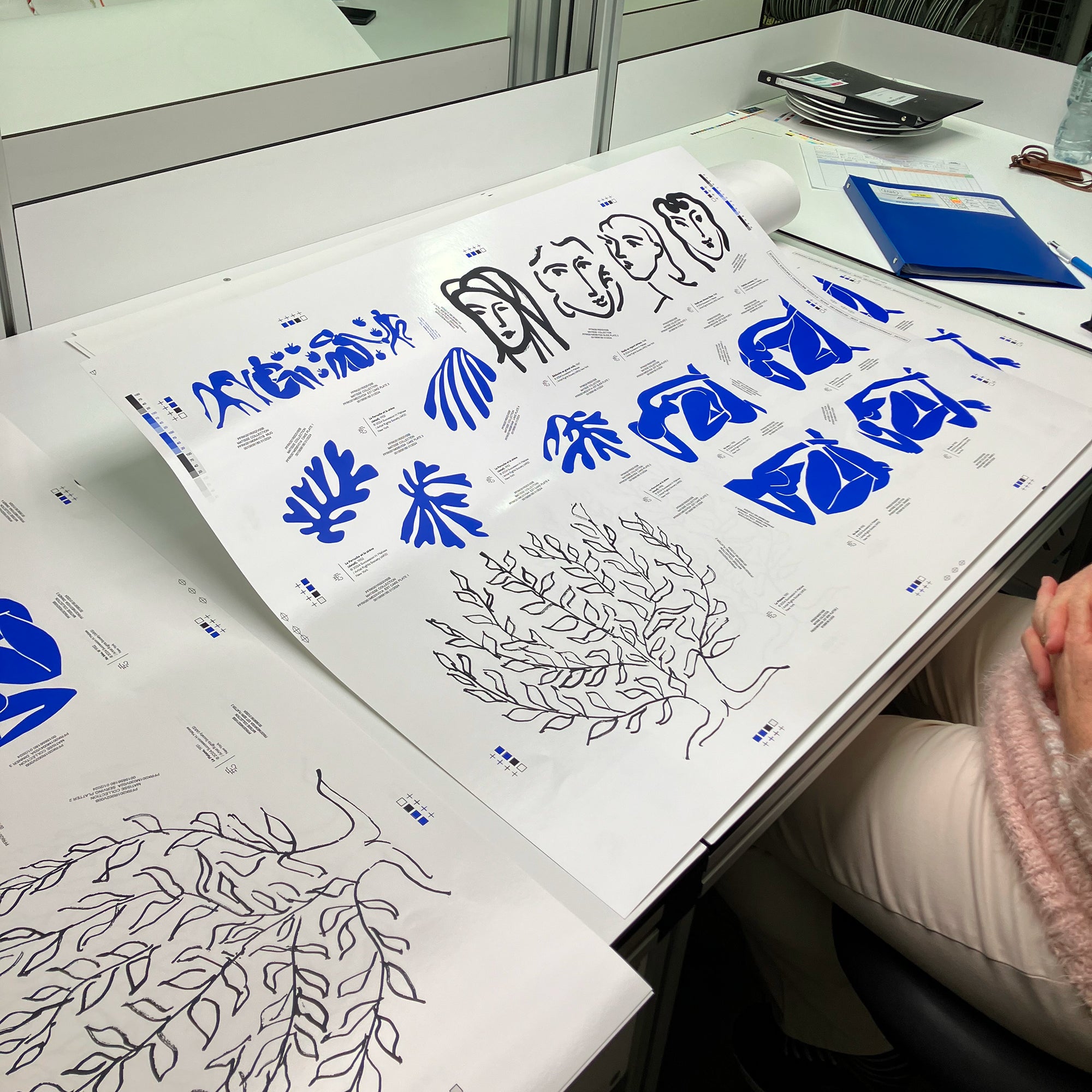 A large printed sheet with blue and black Henri Matisse line drawings spread on a desk, in an office setting.