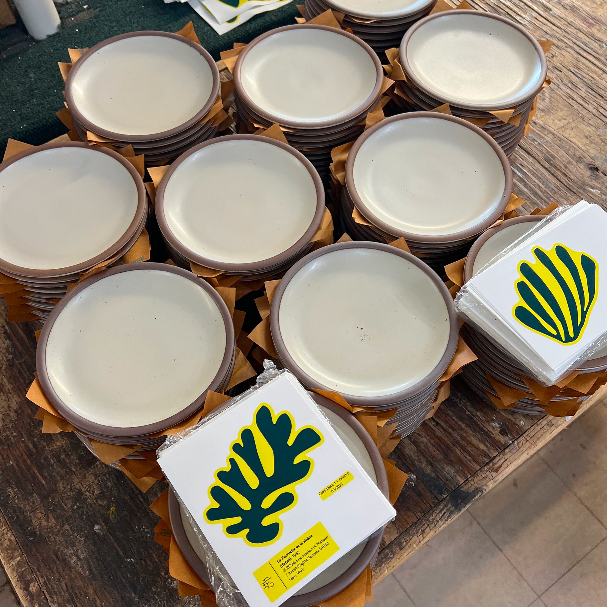 Stacks of small round ceramic plates with off-white centers and brown rims, arranged on a wooden table. Two wrapped stacks show a green and yellow leaf-like graphic on white packaging.