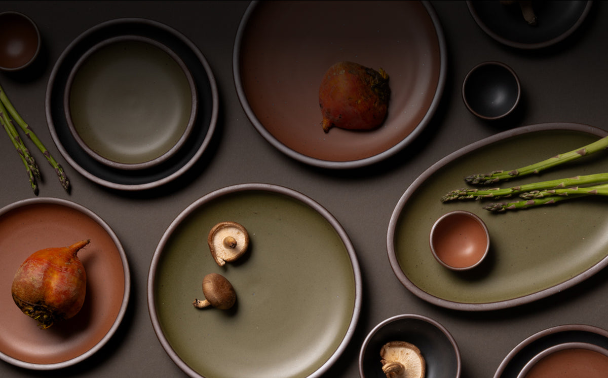 An overhead view of ceramic plates and platters in black, olive, and terracotta colors with food.