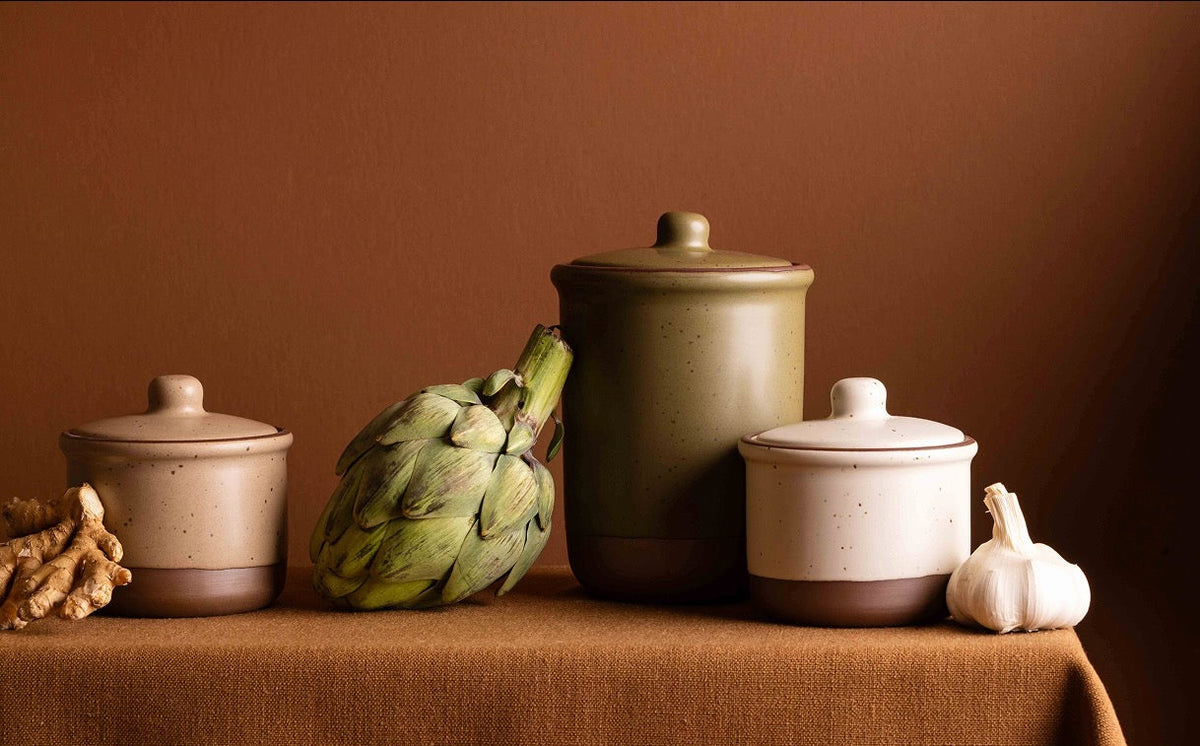 Three ceramic jars with matching lids are arranged on a tablecloth against a warm brown background. The canisters have a two-tone design with a dark brown base and a light beige or cream upper body.