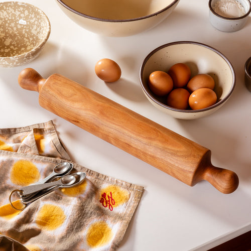 A simple dowel wooden rolling pin with rounded handles, in cherry, on a kitchen counter with measuring spoons, a bowl of eggs, and a hand dyed yellow and brown towel.