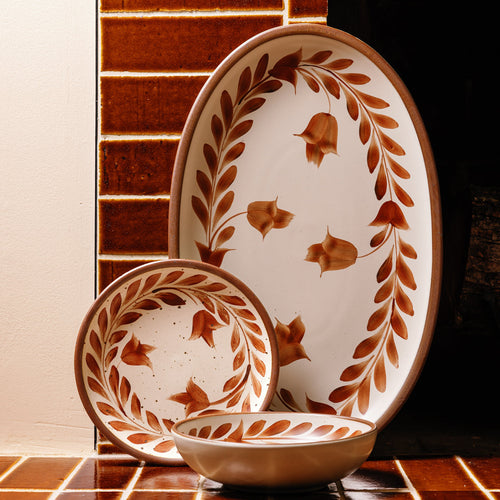 Oval ceramic platter, dinner plate, and everyday bowl with a hand-painted cool terracotta leaf and flower border on a cream background with an exposed clay rim, against a tiled background in a matching color.