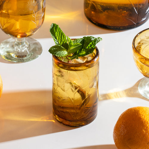 Short amber etched glass tumbler filled with ice, lemon slices, and mint leaf with matching glassware.