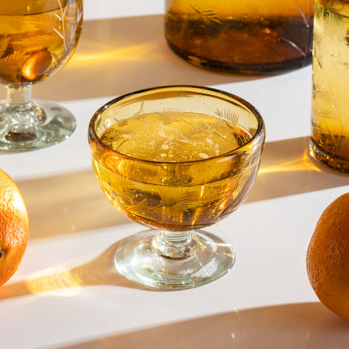 An etched thistle short coupe glass illed with a clear drink next to matching glassware and oranges.