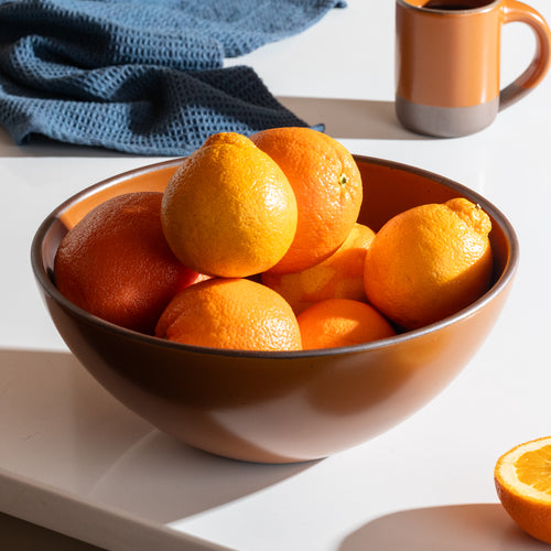 A earthy orange ceramic bowl filled with oranges and lemons on a white table, with a blue textured cloth and an orange mug in the background.