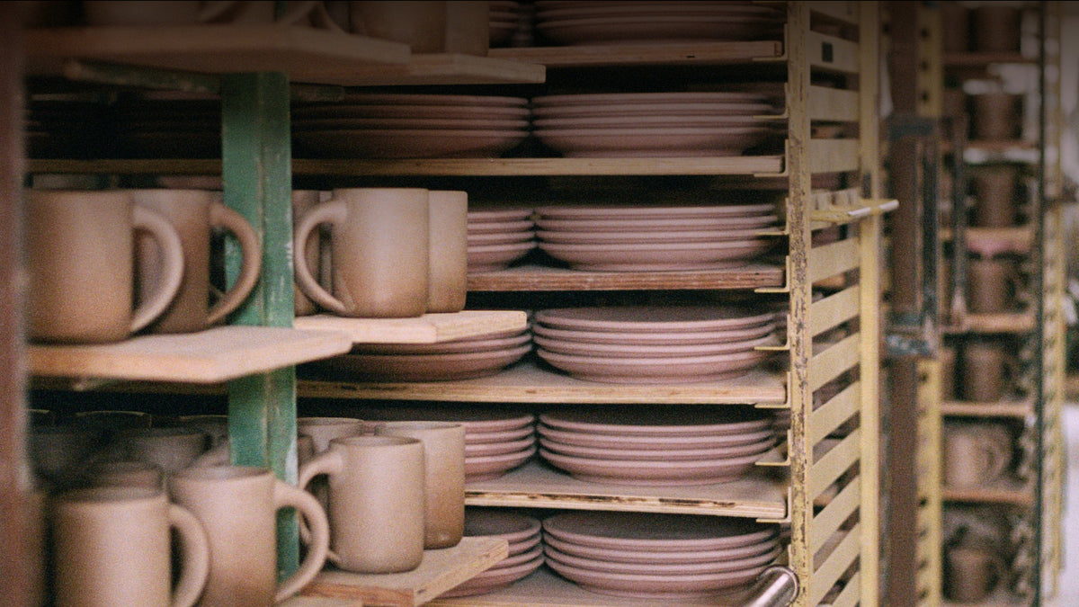 Ceramic bisque mugs and stacked plates displayed on wooden shelves in a pottery kiln storage area.