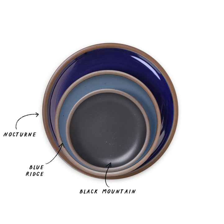 Three nested ceramic plates shown from a top-down view on a white background, with handwritten labels pointing to each: NOCTURNE in the glossy dark blue plate, BLUE RIDGE in a matte denim blue plate,and BLACK MOUNTAIN in a graphite black plate. 