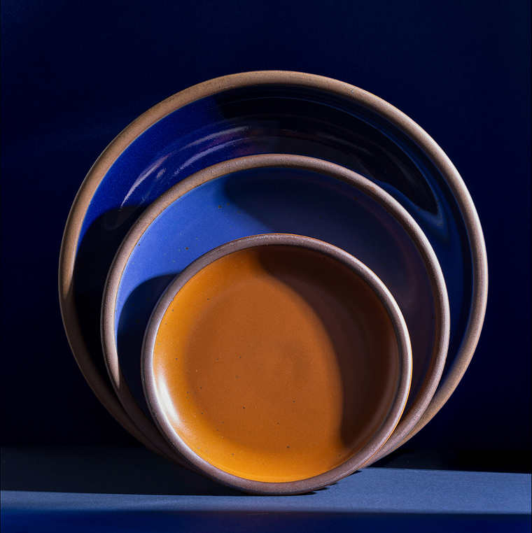 Three nested ceramic plates in a dark glossy blue, a lighter matte blue, and a matte earthy orange, set against a dark blue background.