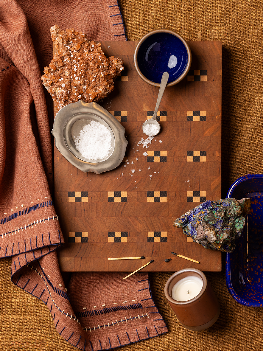 A styled flat-lay of a carved wooden cutting board with geometric inlay patterns, surrounded by earthy props including a terracotta cloth, a dark glossy blue ceramic bowl, a small candle, matches, and mineral-like rocks on a tan fabric background.