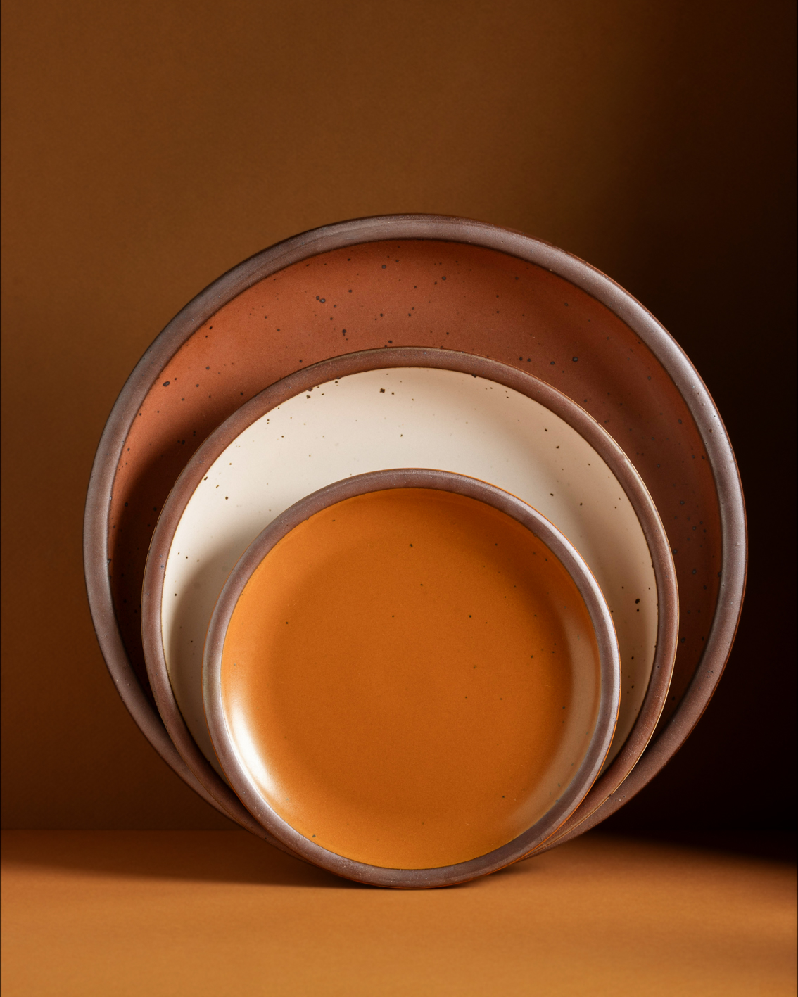 Three nested ceramic plates in a stack, featuring a matte burnt orange dinner plate, a speckled cream side plate, and a glossy burnt orange dessert-sized plate, set against a warm brown background.