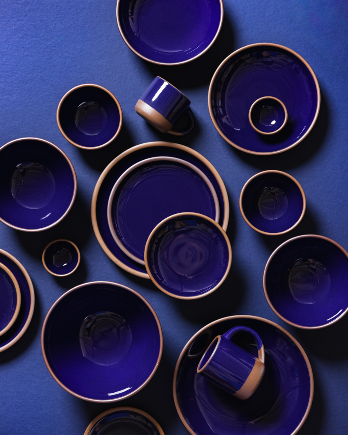 A coordinated set of ceramic tableware in glossy deep cobalt blue with a contrasting light brown rim, arranged in a flat lay on a matching blue background. The set includes plates, bowls, cups, and mugs.