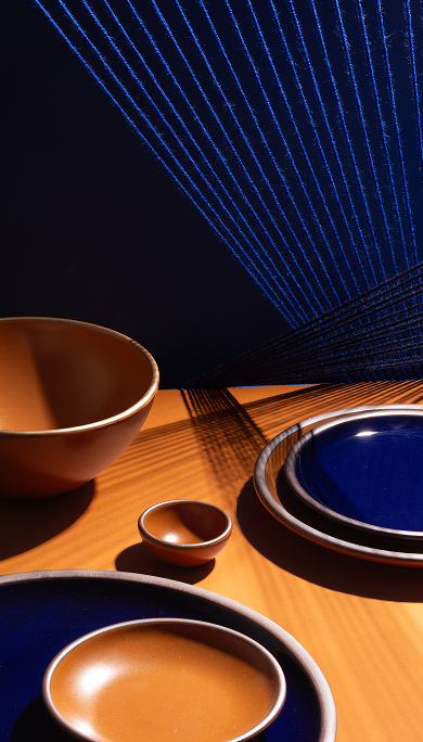 A styled tabletop arrangement of ceramic tableware, including glossy dark blue and earthy orange plates and bowls with light brown rims, set on a warm wood surface under dramatic directional light that casts shadows.