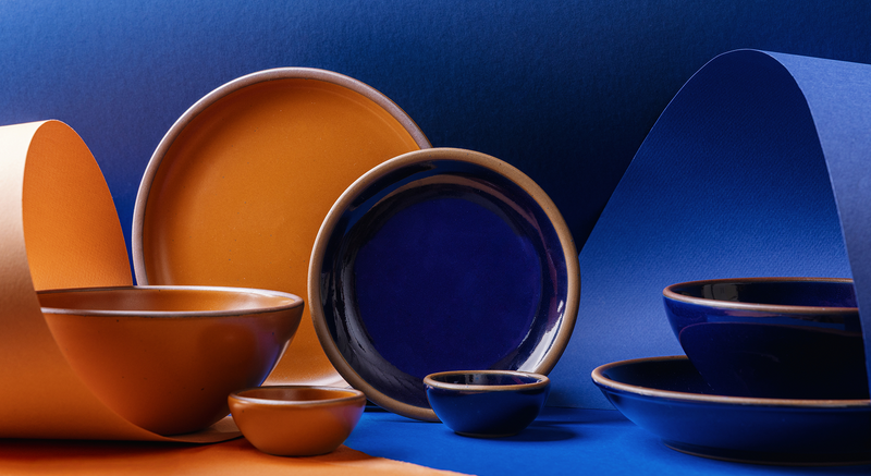 A styled studio arrangement of ceramic tableware in two contrasting glazes: a dark glossy blue and a warm earthy orange. The set includes plates, bowls, and small cups, some nested together and others displayed upright, set against a split blue and orange paper background.